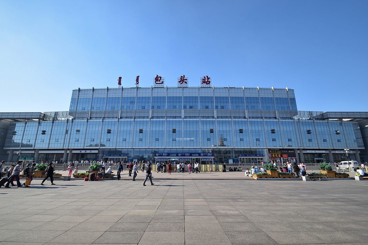 包头站现代站房外观，玻璃幕墙和钢结构屋檐构成横向延展的立面。站前广场开阔，与阿尔丁大街直接相连。