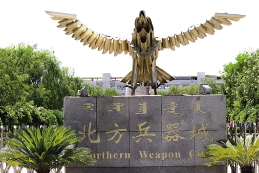 北方兵器城主入口正门，上方有报废炮弹壳焊接成的雄鹰雕塑，下方灰色围墙向两侧延伸，区分展示区与生产区。