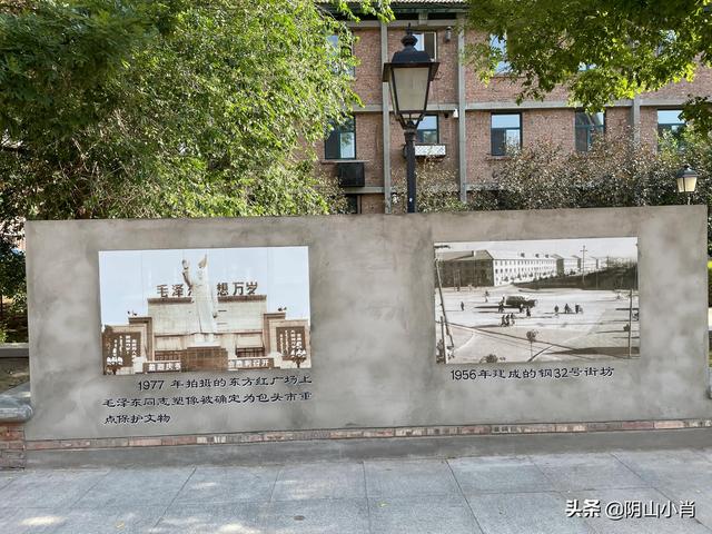 钢32街坊红砖住宅楼前的历史展墙，贴有两幅老照片分别展示1956年建成面貌和1977年东方红广场场景。红砖坡顶楼体本身也清晰可见。
