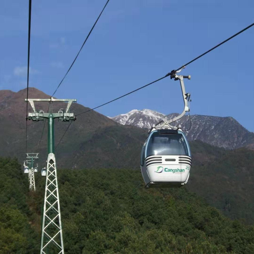 A gondola lift travels over lush green mountains with snow-capped peaks in the background under a cl
