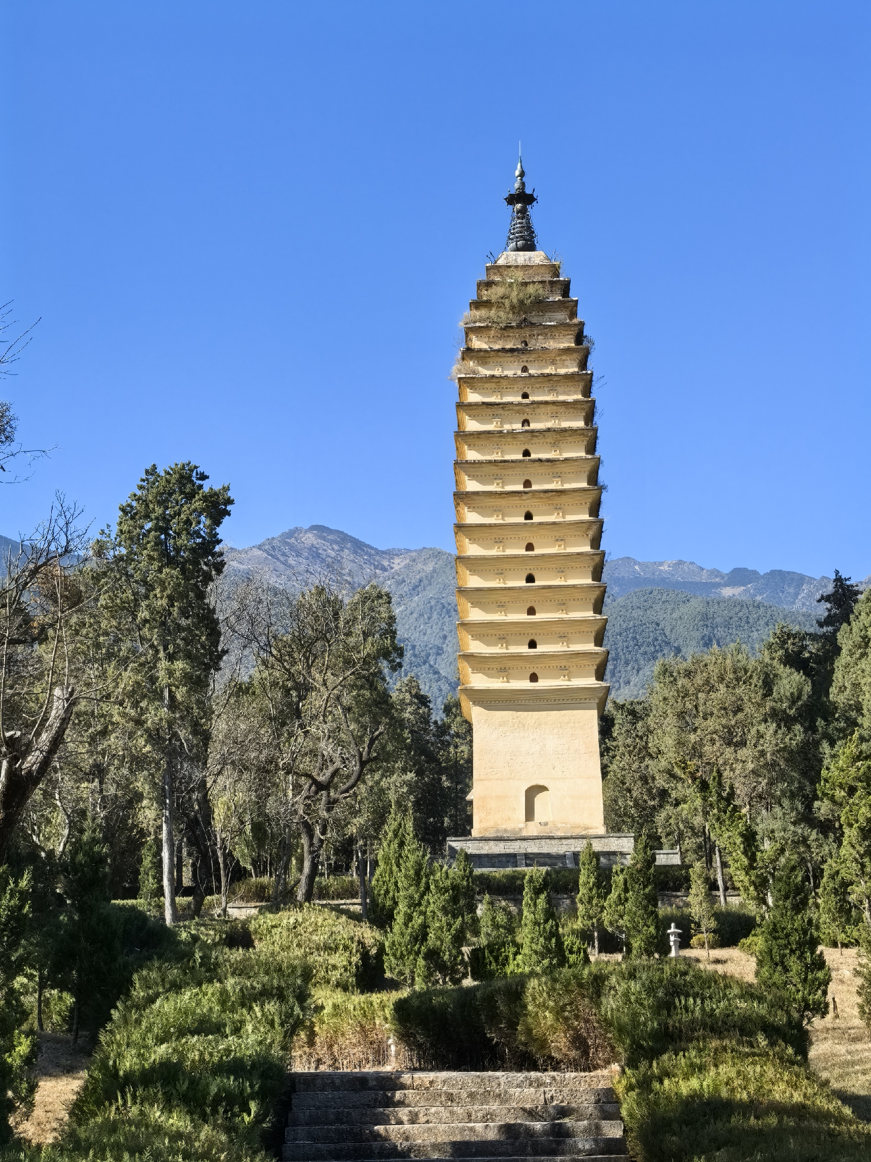 A tall, ancient Buddhist pagoda with multiple tiers rises amidst trees, with mountains in the backgr