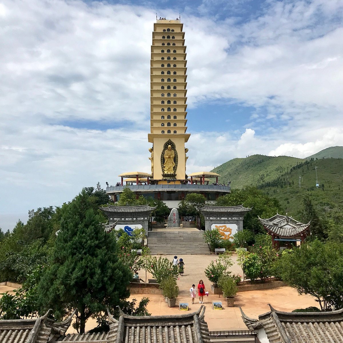 A tall, multi-tiered pagoda with a golden Buddha statue at its base stands prominently amidst lush g