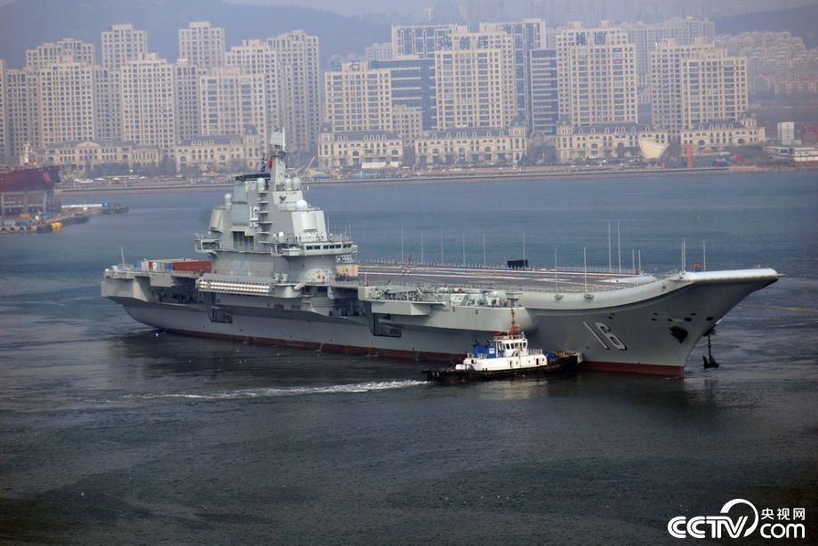 A large aircraft carrier, likely the Liaoning, is being moved through a harbor b