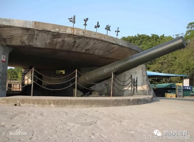 A large artillery cannon is displayed outdoors near a fortress with signs on a concrete platform, overlooking the bay at