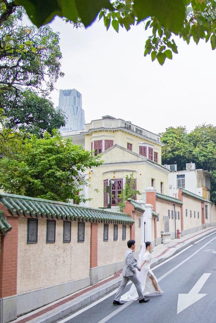 A newlywed couple walks along a street with a colorful building in the background, surrounded by lush greenery and tradi