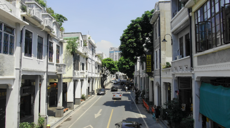 A street lined with white, layered colonial-style buildings with balconies and terrace walkways, shaded by trees under a