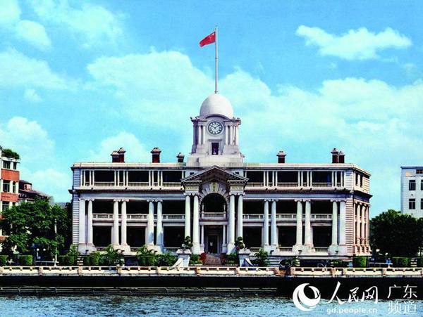 A historic clock tower with a flagpole atop, situated along a riverbank, featuring classical architectural details and a