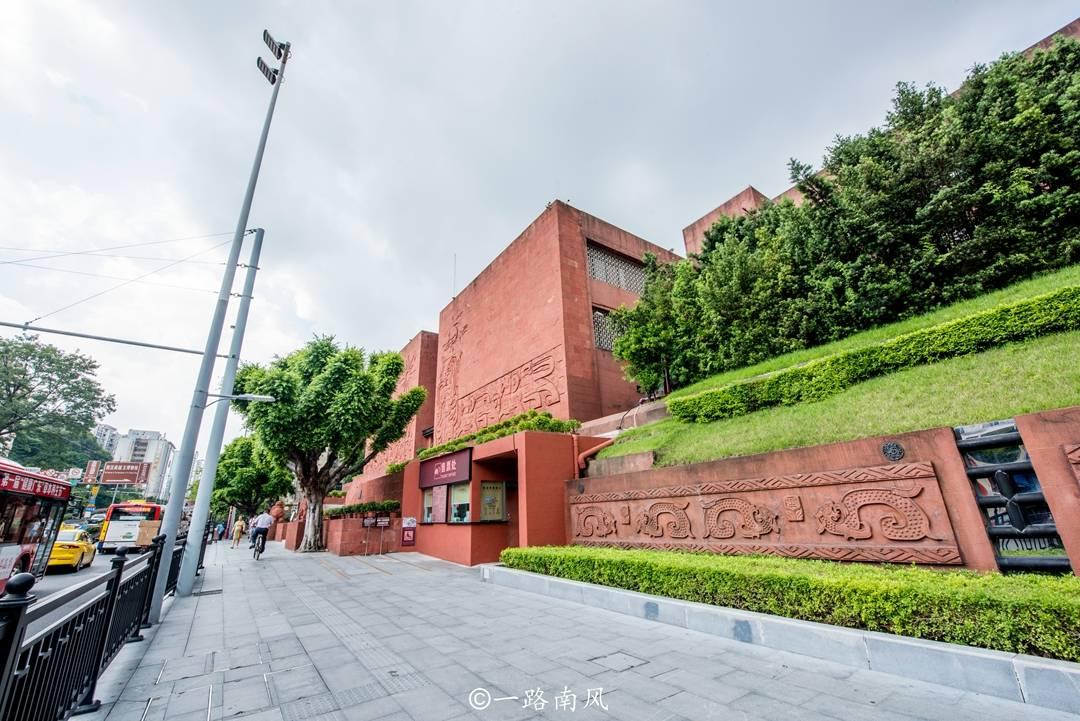 A red-brick building with carved patterns and greenery surrounds the entrance, situated along a sidewalk in an urban are