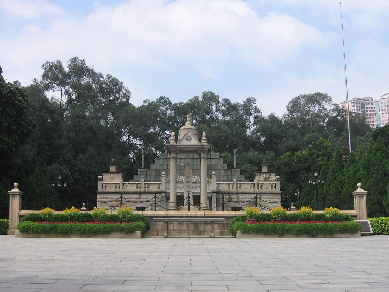 A memorial monument surrounded by lush greenery, with an ornate structure at its center, set against a backdrop of trees