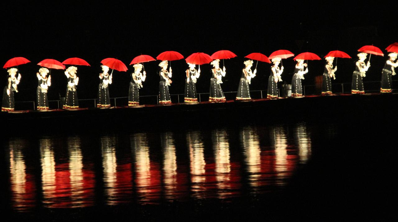 夜间演出全景：演员在竹排上高举红色雨伞，远山与水面在灯光下融为一体