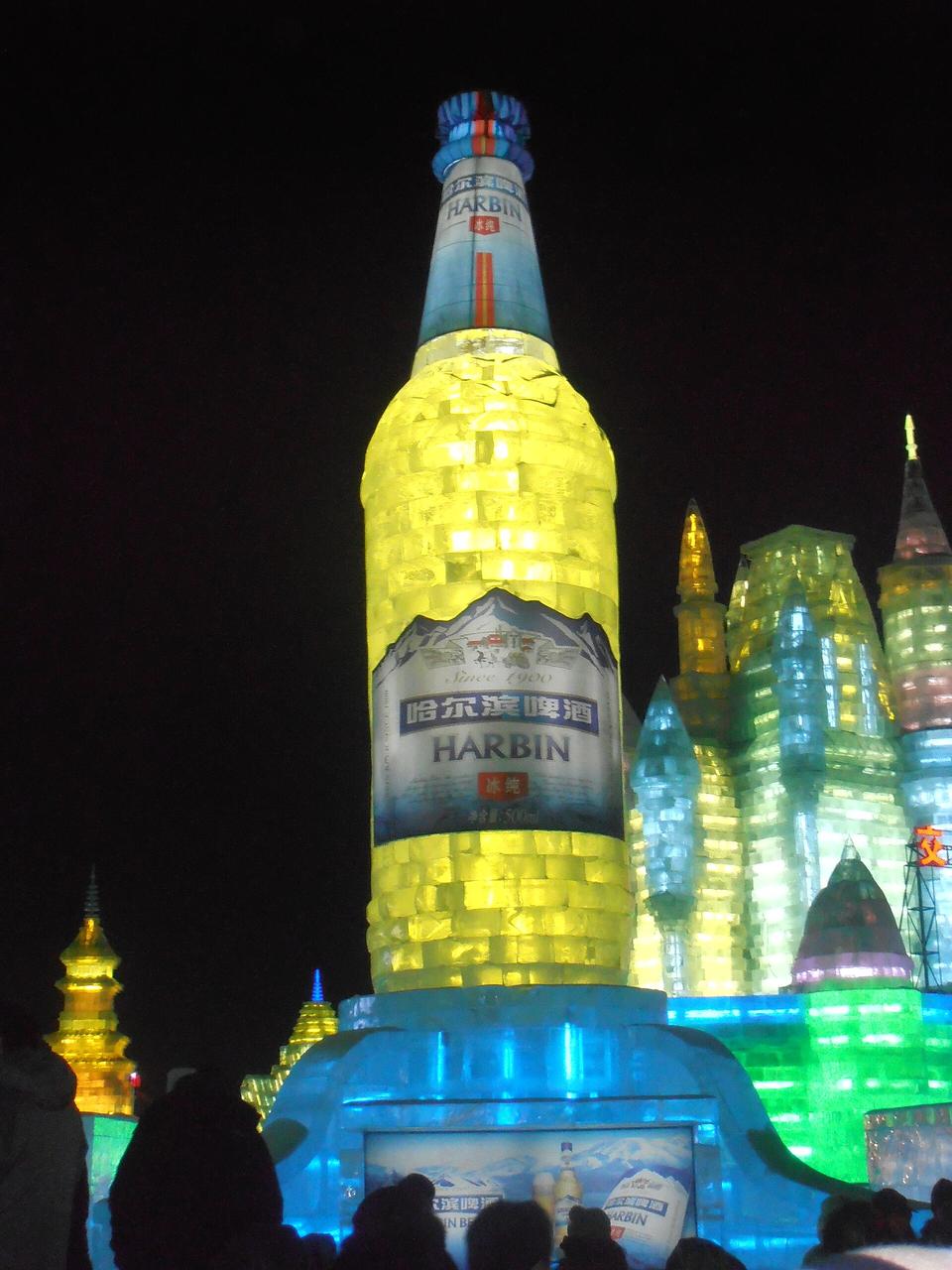哈尔滨冰雪大世界内的巨型啤酒瓶冰雕，夜晚彩色灯光照射下格外醒目，周围是冰雪城堡造型