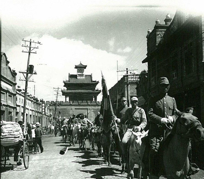 A line of soldiers in uniform on horseback and foot, carrying flags, is passing 