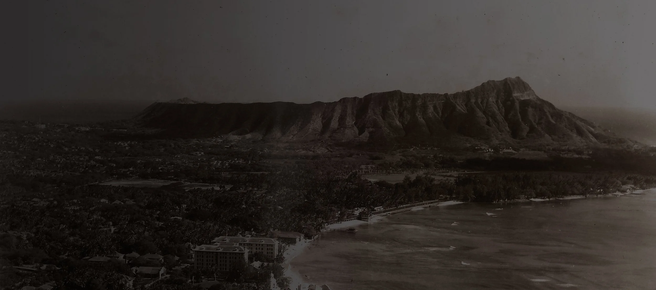 Waikīkī Historic Trail 的冲浪板形标识牌位于人群行走的人行道上，显示牌子高度、bronze 材质与周围旅游酒店环境的关系