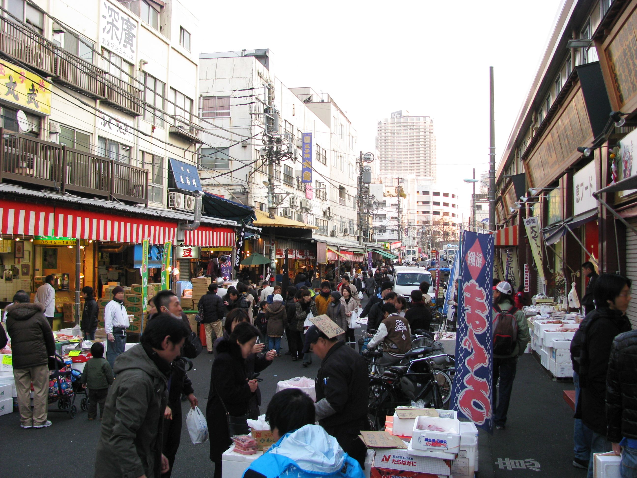 筑地场外市场的窄巷，两侧商店招牌密集排列，行人与运货车交错