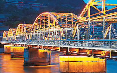 A brightly illuminated roller coaster structure extends over a river with reflec