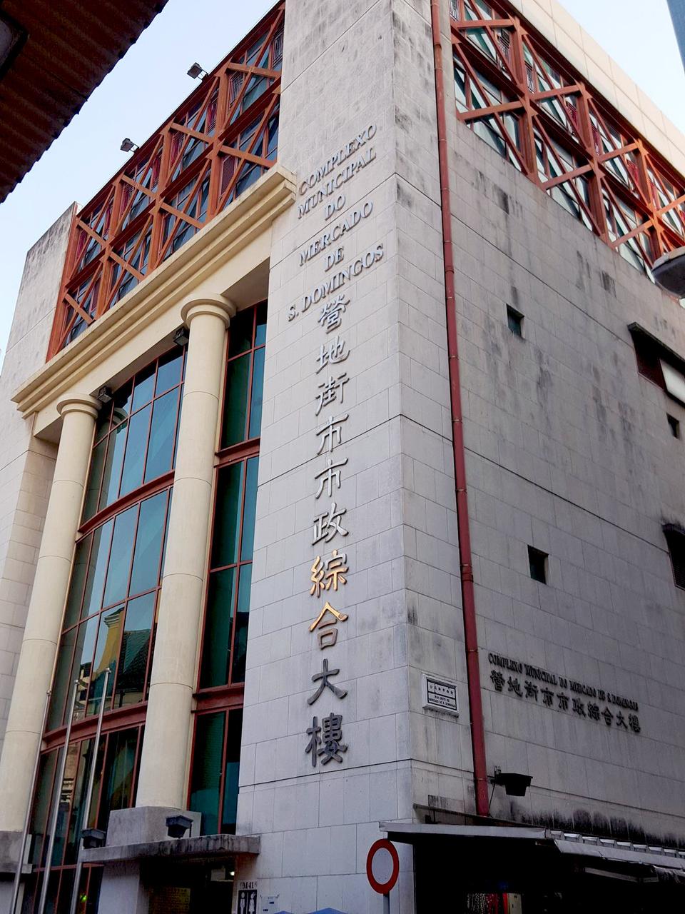 营地街市外立面：七层米黄色现代建筑，红色窗框，地面层为摊档
