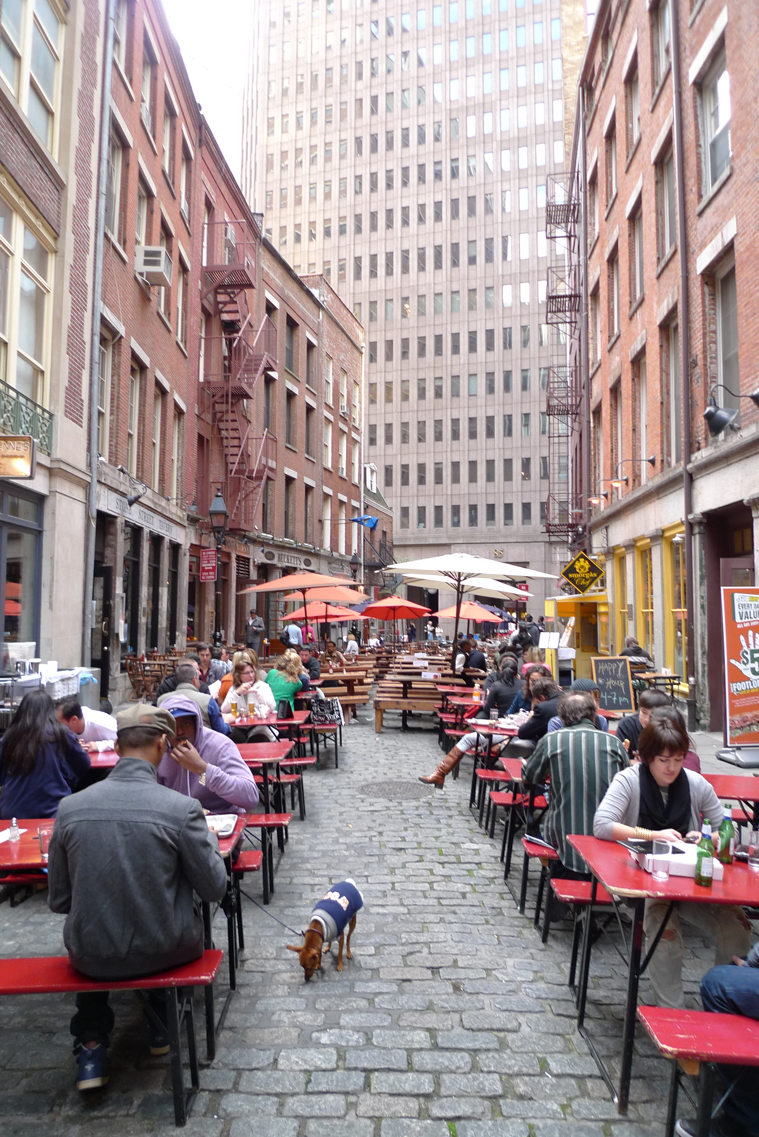 Stone Street 东段，中间是比利时块石铺装和户外餐桌，两侧是灾后重建砖楼，背景可见 85 Broad Street 大厦