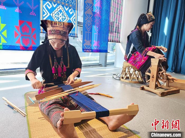 传习所教学区场景,学员在老织工指导下练习腰织机操作,墙上可见传承人简介展板,男性学员也在其中