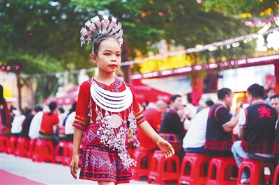 保亭七仙文化广场上，身着黎族服饰的儿童在红毯上走秀，红色塑料凳观众席坐满人群，背景为节庆装饰和绿树