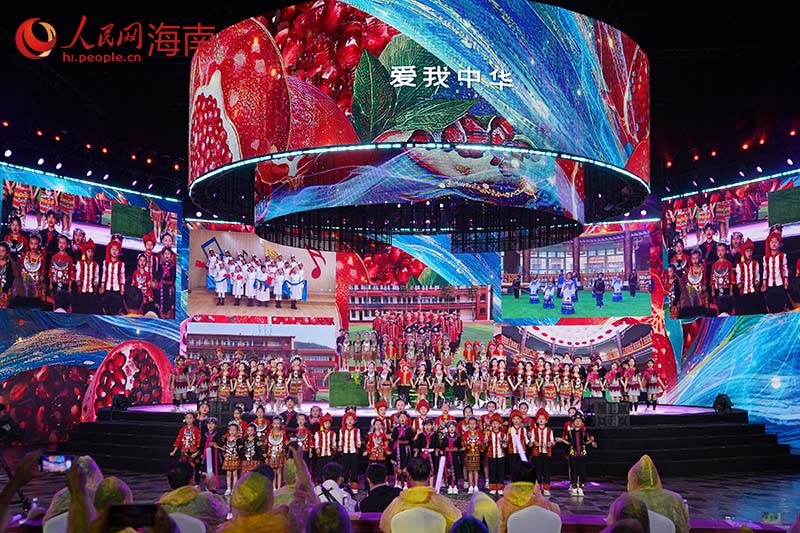 五指山市三月三广场圆形LED环形屏下，身着民族服装的儿童在阶梯舞台上合唱，"爱我中华"字样亮于环屏
