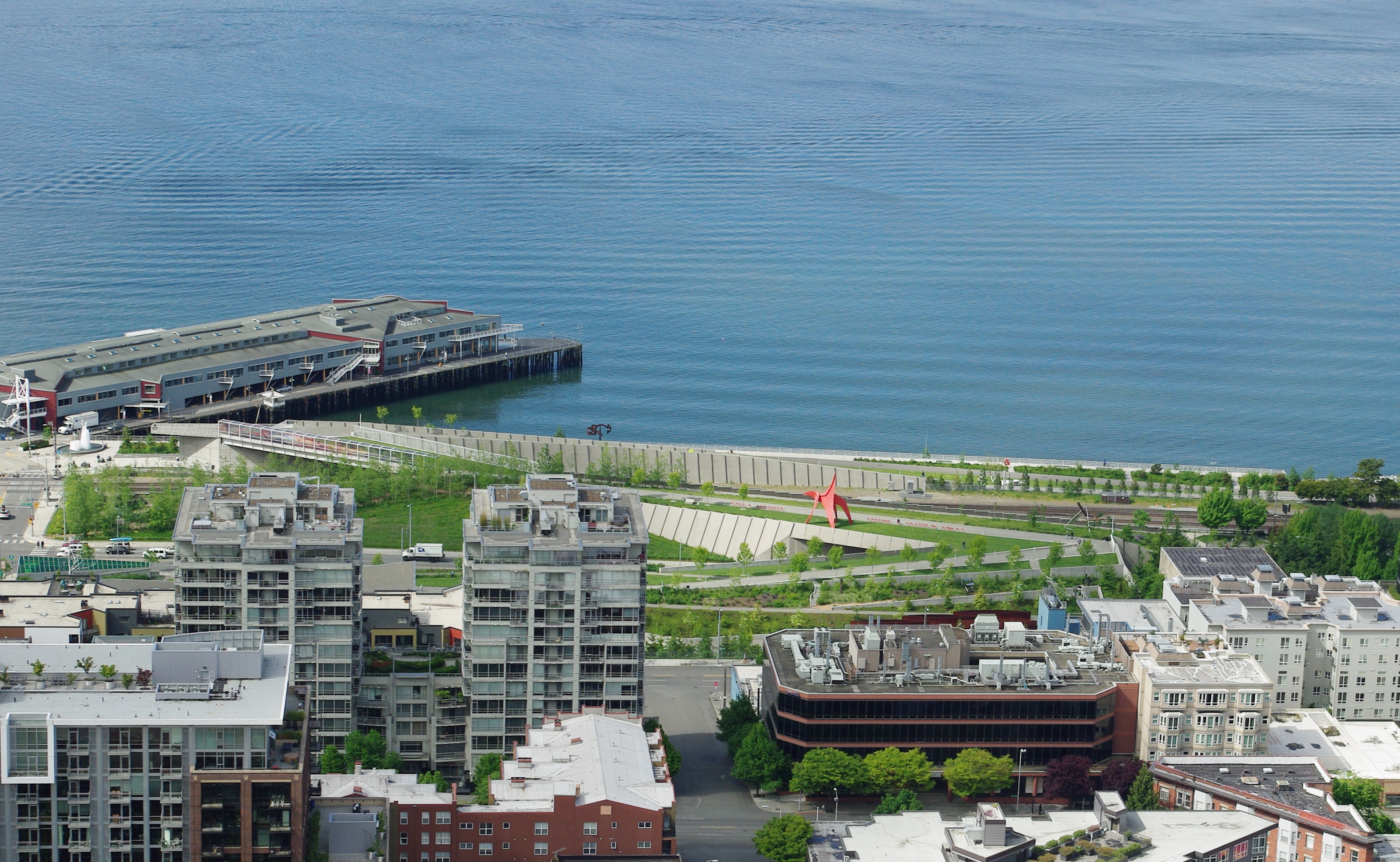 从 Space Needle 高位俯拍的 Olympic Sculpture Park：Z 形平台从城市端的 PACCAR Pavilion 跌落到 Elliott Bay 水边，跨过 Elliott Avenue 与 BNSF 铁路两道线性基础设施