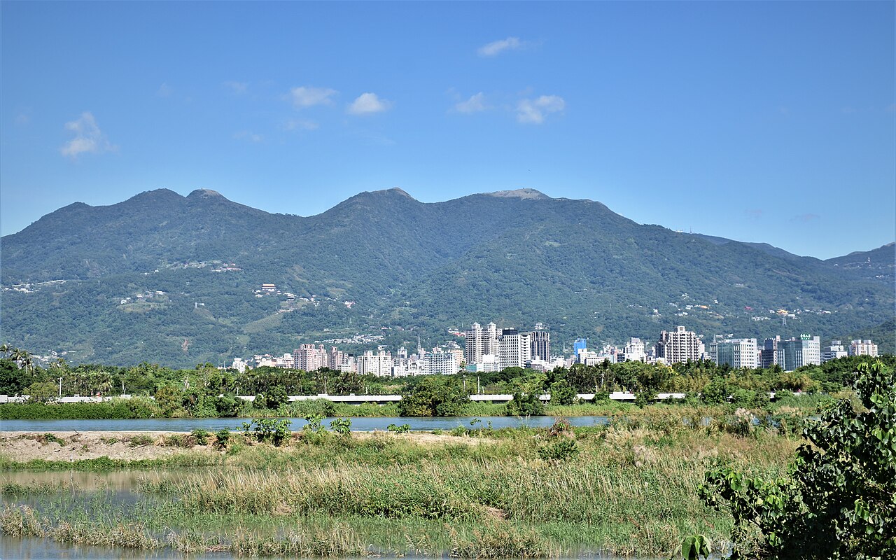 A city skyline lies below lush mountains with a blue sky overhead, across a wetl