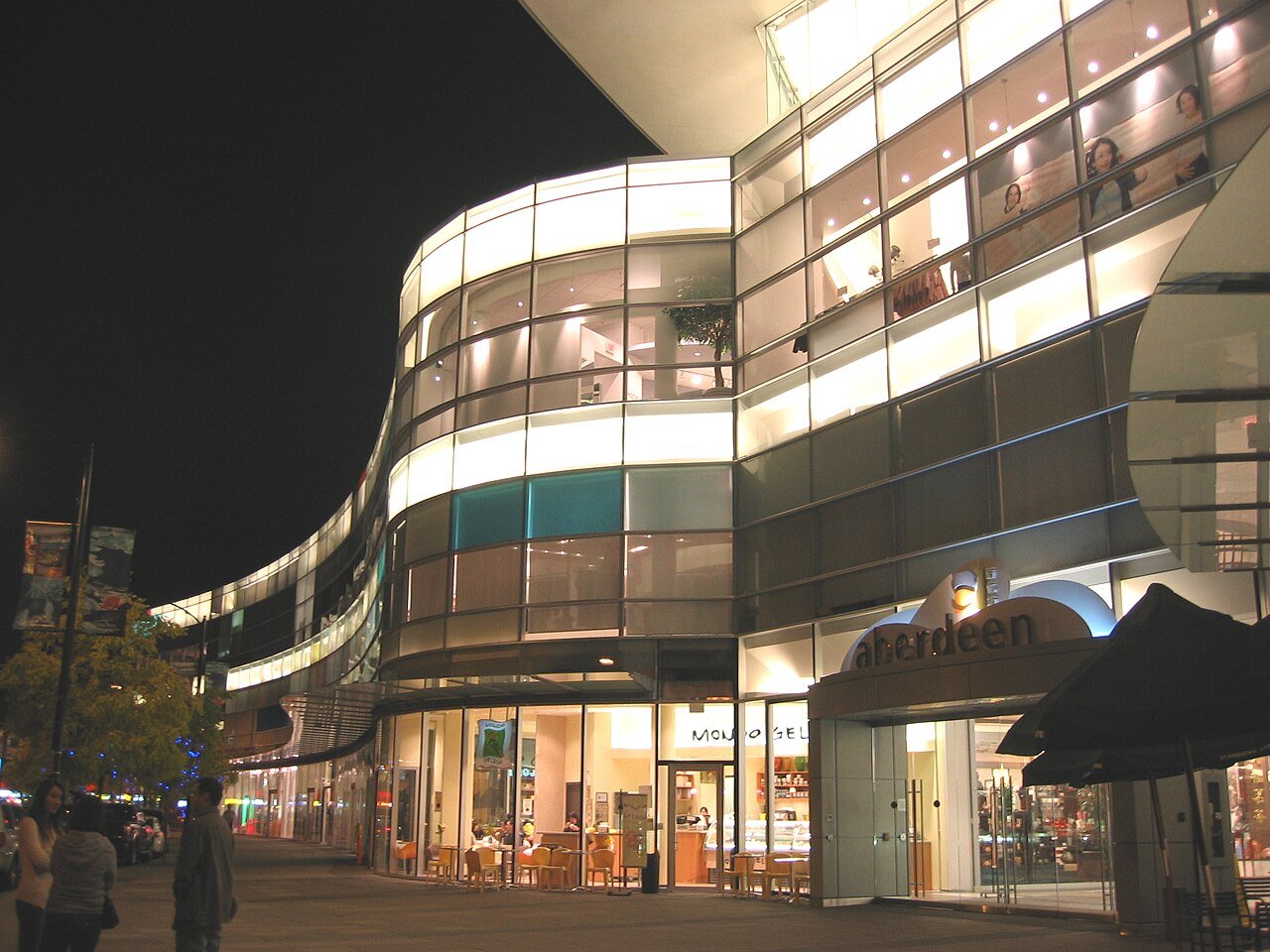 Aberdeen Centre 夜间玻璃外立面，弯曲幕墙和多层室内灯光从 Hazelbridge Way 一侧可见