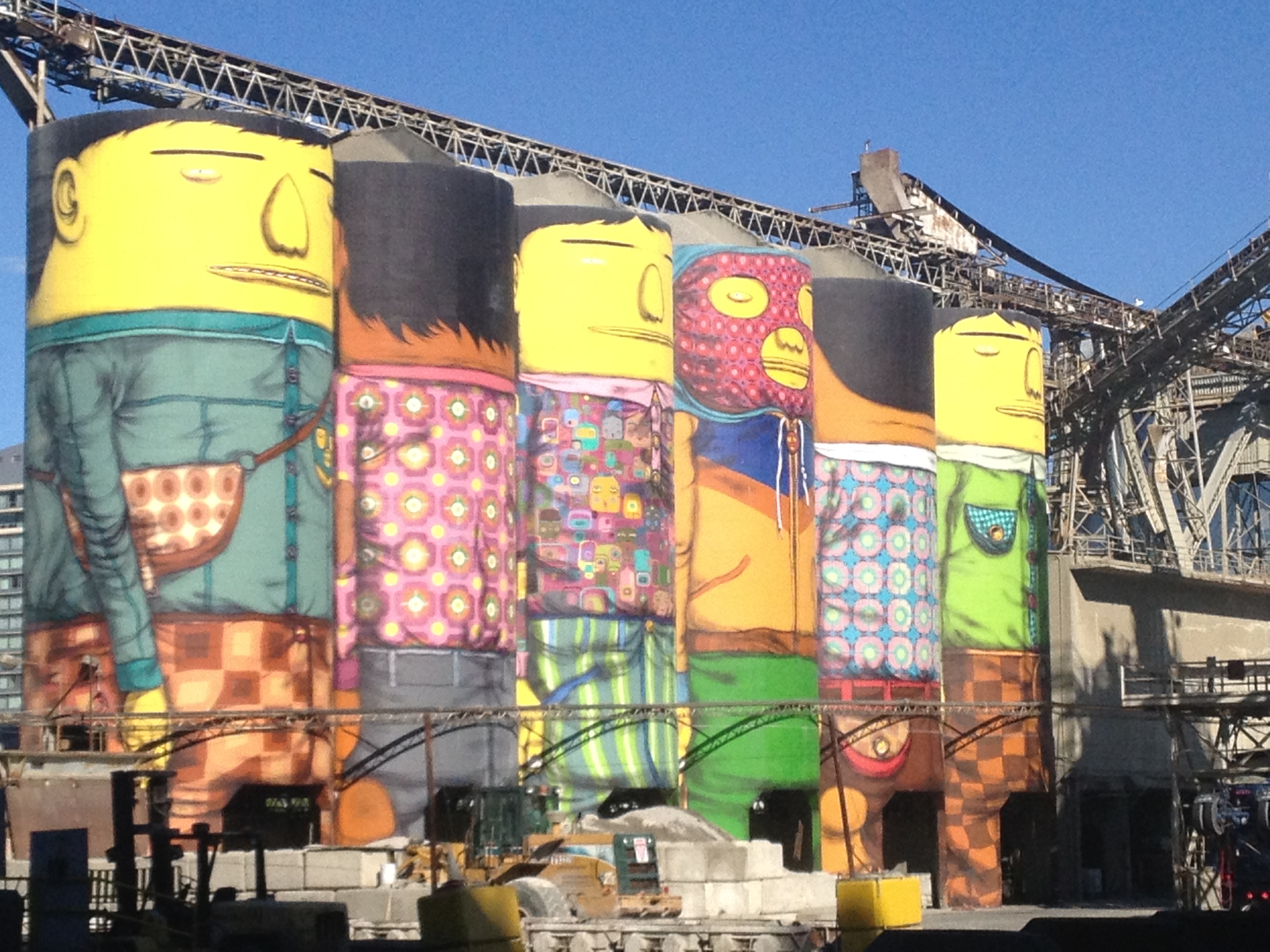 OSGEMEOS Giants 彩绘壁画覆盖 Ocean Cement 六座 21.3 米高水泥仓筒全景，仓筒紧邻 False Creek 水面，从 Granville Street Bridge 上拍摄
