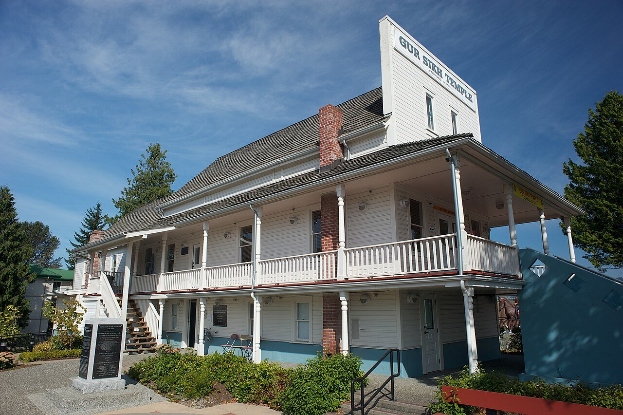 Gur Sikh Temple 正面外观，false-front 木结构临街立面，木 siding、山墙屋顶被平整假墙遮蔽，门前草地和右侧 Nishan Sahib 旗杆