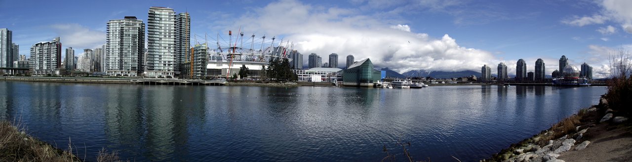 Habitat Island 全景：卵石滩环绕的人工岛，植被密集，背景为 Olympic Village 住宅楼和 False Creek 城市天际线