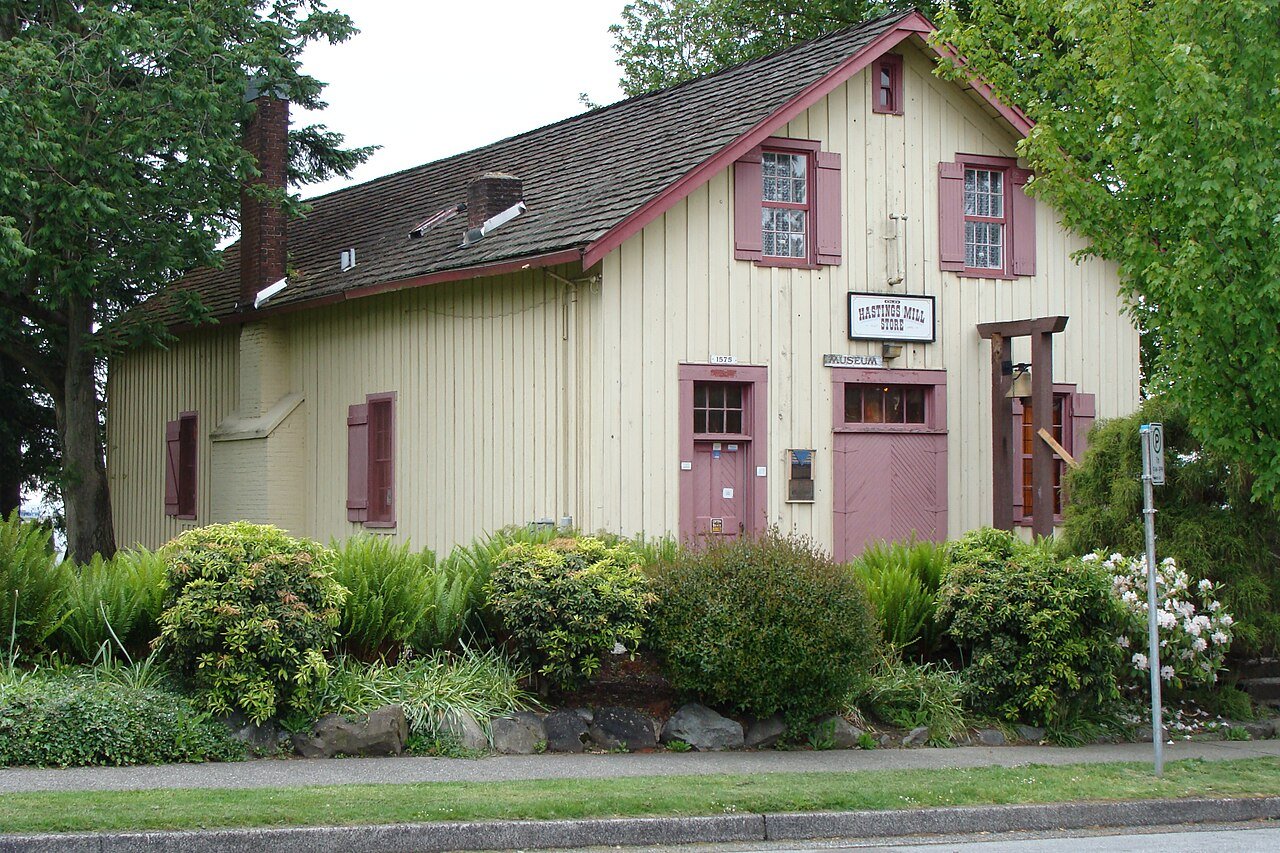 Hastings Mill Store Museum 正面，两层清水木墙建筑，门廊和窗户可见，草坪上立着铜钟