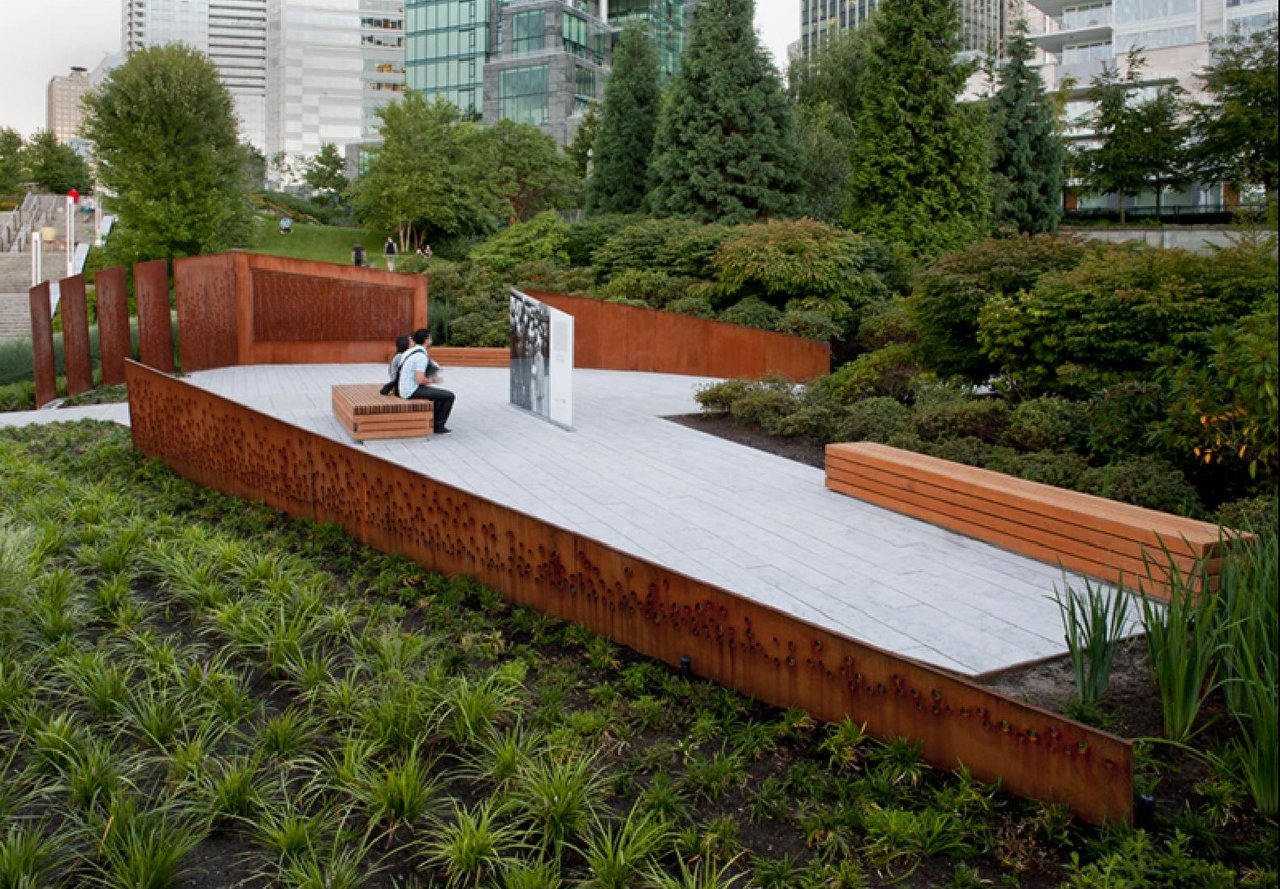 Komagata Maru Memorial 全景：锈色穿孔钢板、中央玻璃面板和公共长凳从右至左排列，背景是 Coal Harbour 水面