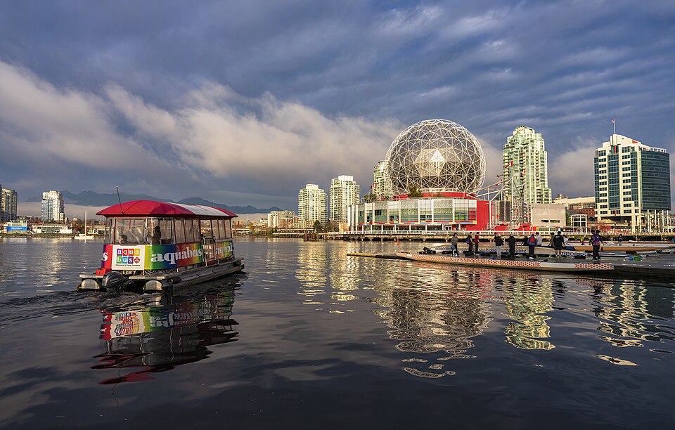 从 False Creek 水面看穹顶与北岸玻璃住宅塔楼，画面中穹顶矮、半透明，右侧高层方正不透明