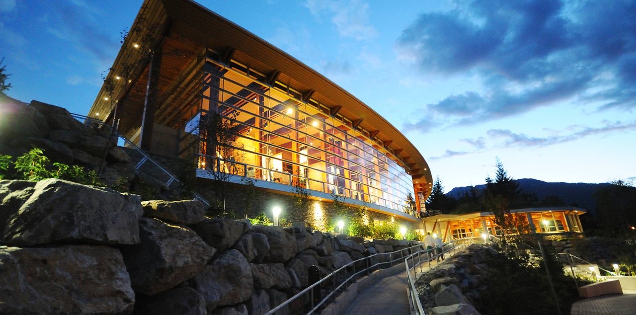 SLCC 外部黄昏景观，长向 longhouse 屋脊与弧形 istken 体量并置在同一栋建筑上，面向 Blackcomb Way 和山地森林