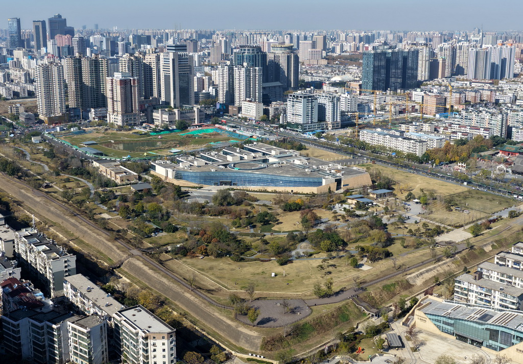 从空中俯瞰郑州商城遗址公园，城墙残段沿公园绿带延伸，后方是密集的城市高层建筑